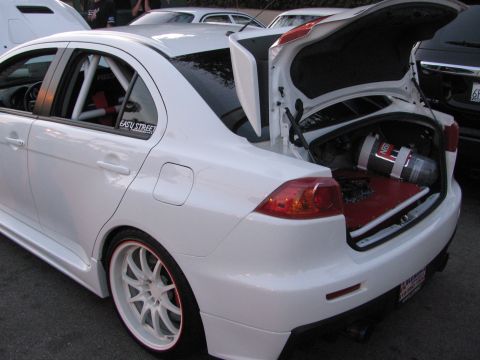 1990 Ford Bronco - white Mitsubishi Lancer Evolution on white wheels with its trunk open, revealing a mounted fuel cell, at a car show.