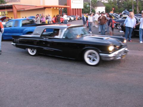 1990 Ford Bronco - black 1950s-era convertible with chrome trim and whitewall tires rolls past spectators at a dusk car show.