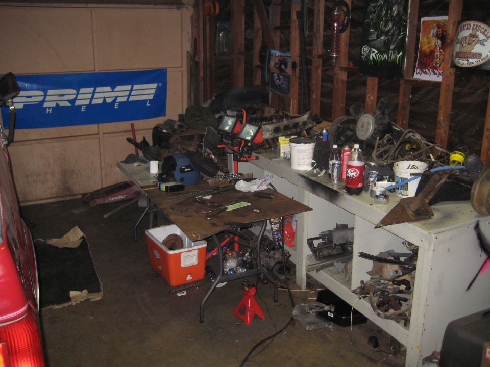 Garage interior with workbench cluttered with engine parts and tools. Red car visible on left