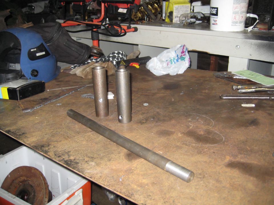 Metal rod and two cylindrical parts on a workbench with tools and a welding mask in the background
