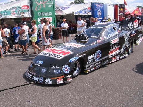 1998 GMC Sierra 1500 - A black dragster with sponsor decals is parked on a paved surface, with people and tents visible behind it.