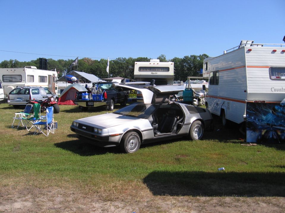Silver DeLorean DMC-12 with gullwing doors open, parked on grass next to RVs
