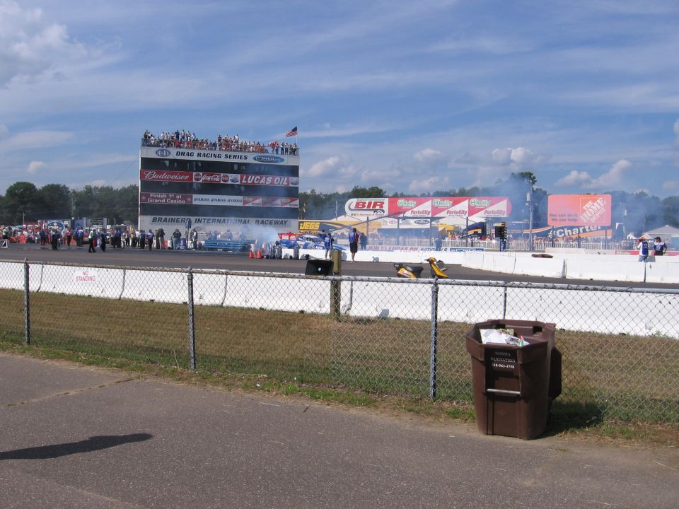 Drag strip with race cars, spectators, and a large scoreboard tower. Smoke is coming from the track