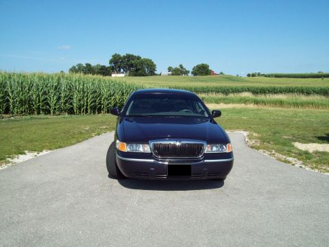 1999 Mercury Grand Marquis - Head-on front view of a dark navy blue Mercury Grand Marquis sedan on a paved rural drive, showing its chrome waterfall grille with the Mer…