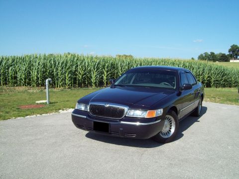 1999 Mercury Grand Marquis - Dark navy blue Mercury Grand Marquis sedan parked on pavement in front of a tall green cornfield under a clear blue sky, shot from a low fr…