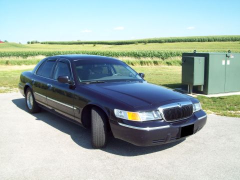 1999 Mercury Grand Marquis - Front-quarter view of a dark Mercury Grand Marquis sedan parked on a road shoulder beside a green cornfield.