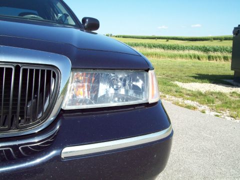 1999 Mercury Grand Marquis - Close-up of the passenger-side front corner of a dark navy blue Mercury sedan, showing the clear composite headlight with amber turn-signal…