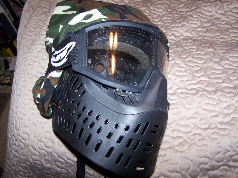 Black paintball mask with camouflage fabric and clear goggles rests on a quilted surface