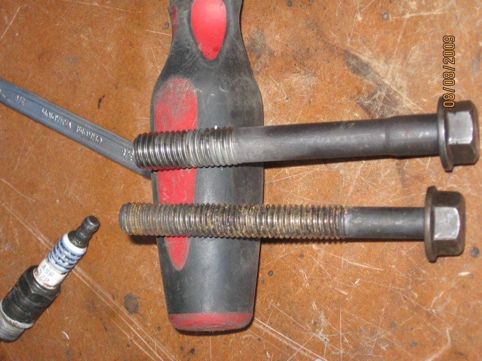 Two bolts and a spark plug on a wooden surface, with a wrench and screwdriver handle visible