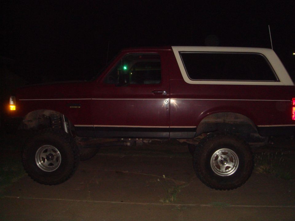 Maroon 1992 Ford Bronco with white top, lifted suspension, and aftermarket wheels, seen from the side at night