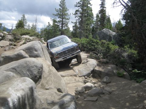 1986 Ford Bronco - A blue Ford Bronco is shown traversing a challenging rocky terrain with dense foliage in the background.