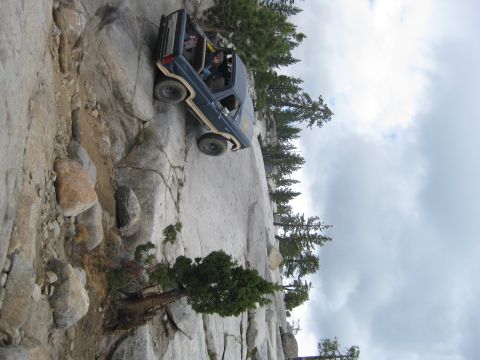 1986 Ford Bronco - A dark blue SUV navigates a challenging rocky terrain, with trees and a cloudy sky overhead.