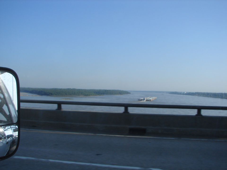 View from a bridge of a wide river with barges, showing a side mirror reflection of a vehicle