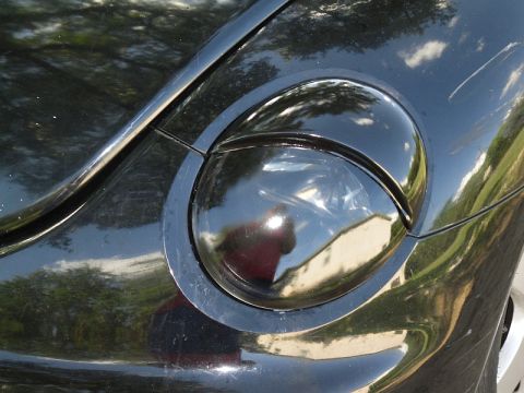1999 Volkswagen Beetle - Close-up of a black car's headlight assembly, showing reflections of the surrounding environment.