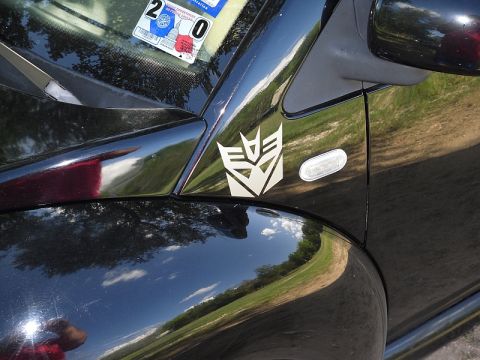 1999 Volkswagen Beetle - Black car fender and door with Autobot logo sticker. Reflection of trees and sky.