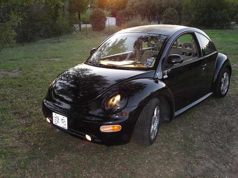 Black Volkswagen Beetle parked on grass with trees in background. Has a Transformers Autobot logo on fender