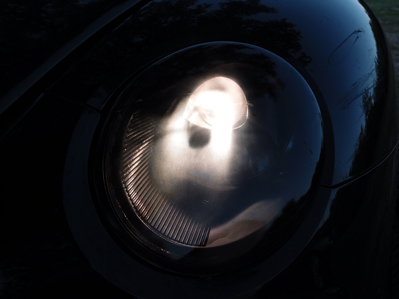 Black 1999 Volkswagen Beetle headlight with the bulb illuminated, showing ribbed glass and a curved body panel