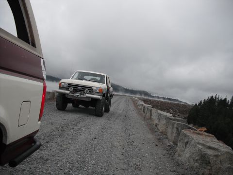 1991 Ford Bronco - Two off-road vehicles are on a gravel road with a stone barrier and misty mountains in the background.