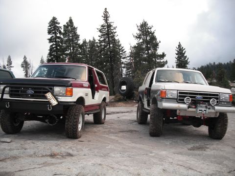 1991 Ford Bronco - Two modified Ford Broncos are parked side-by-side on a rocky terrain, with a forest visible behind them.