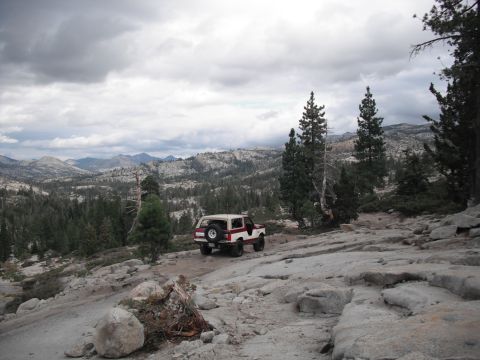 1991 Ford Bronco - A two-tone SUV navigates a challenging rocky path with a vast, forested mountain landscape under a cloudy sky.