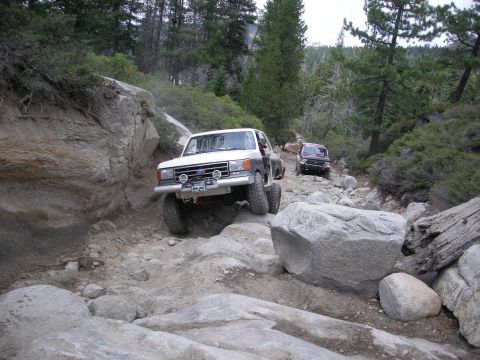 1991 Ford Bronco - Two SUVs tackle a challenging off-road path with large rocks and steep inclines.