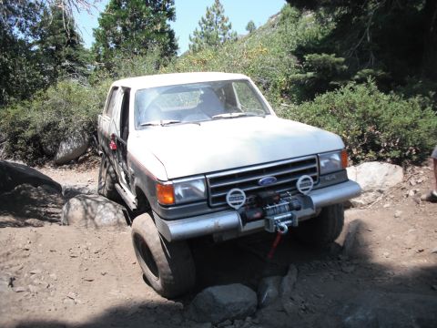 1991 Ford Bronco - This Ford Bronco is shown off-roading on a rocky trail with trees and bushes in the background.