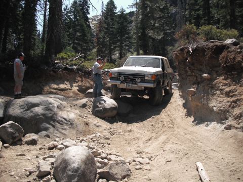 1991 Ford Bronco - An off-road vehicle carefully maneuvers through a challenging, rocky terrain with dense forest surroundings.