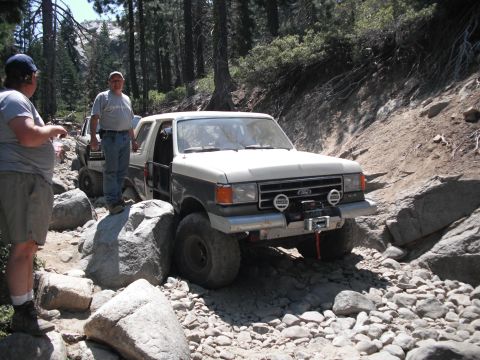 1991 Ford Bronco - Two Ford Broncos navigate a challenging, rocky off-road trail in a wooded area.