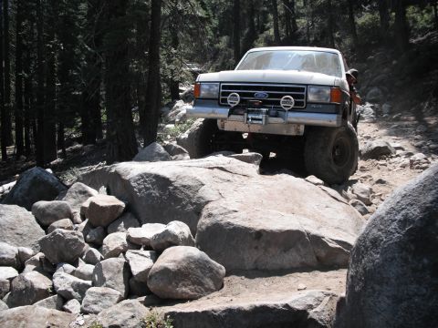 1991 Ford Bronco - A lifted Ford Bronco navigates a challenging, rocky off-road path through a forest.