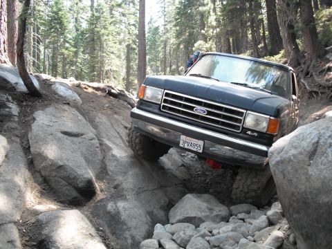 1991 Ford Bronco - A dark-colored Ford truck is shown off-roading on a steep, rocky path surrounded by trees.