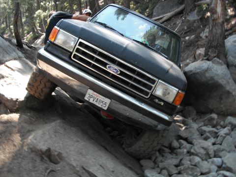 1991 Ford Bronco - A black Ford Bronco is shown off-roading on a rocky trail, with its front passenger side tire elevated.