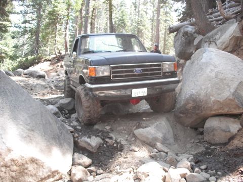 1991 Ford Bronco - This Ford truck is shown on a challenging off-road trail surrounded by trees and large rocks.