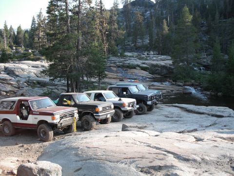 1991 Ford Bronco - A lineup of four off-road SUVs, including a 1991 Ford Bronco, parked on a rocky bank beside a flowing stream.
