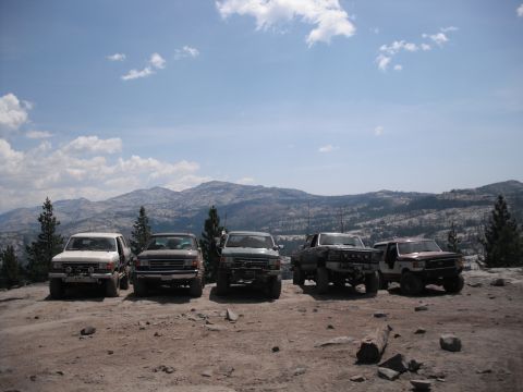 1991 Ford Bronco - A group of off-road trucks are parked on a rocky terrain with a mountainous landscape under a partly cloudy sky.