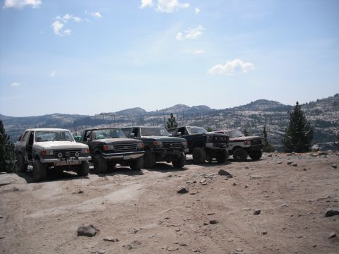 1991 Ford Bronco - Several off-road vehicles are parked in a line on a dusty mountain road under a partly cloudy sky.