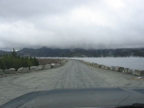 1989 Ford Bronco - View from inside a vehicle driving on a gravel road towards a dam, with misty mountains in the distance.