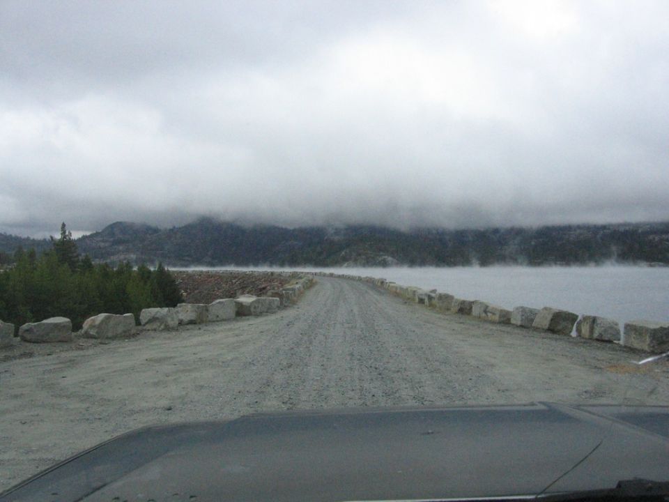 Dark dashboard in foreground, gravel road leading to a dam with mountains and fog in background