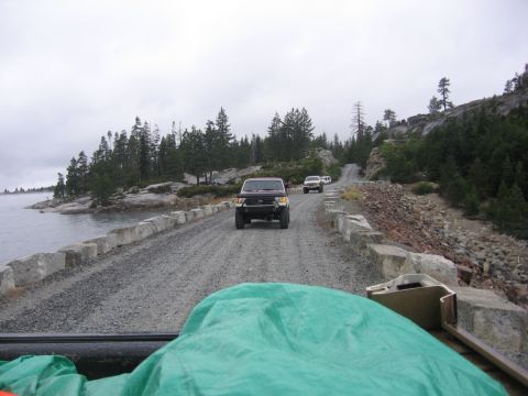 1989 Ford Bronco - Multiple SUVs travel on a gravel road along a lake, with trees and rocky terrain visible.
