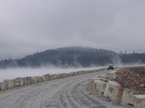 1989 Ford Bronco - A dark SUV drives down a gravel road bordered by a stone wall, with fog obscuring the distant trees and hills.