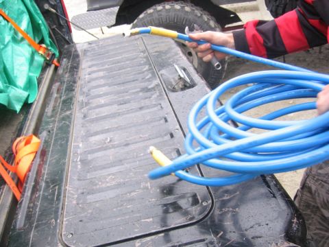 1989 Ford Bronco - A person is holding a blue hose in the bed of a truck, which also contains orange ratchet straps and a green tarp.
