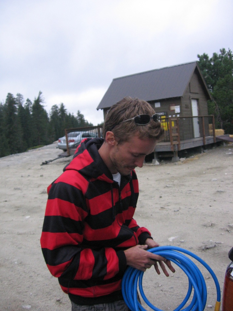 Man in a red and black striped hoodie holds a coiled blue hose. Small cabin in background