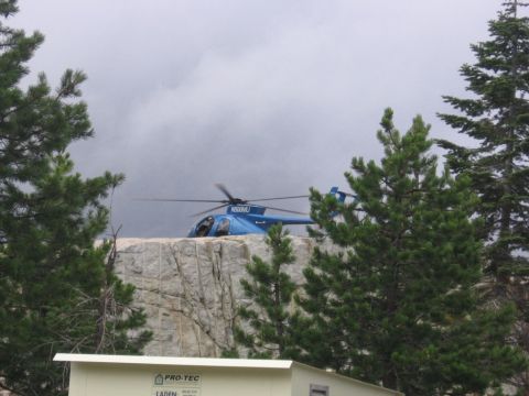 1989 Ford Bronco - A blue helicopter lands on a rocky outcrop, with trees framing the scene under a cloudy sky.