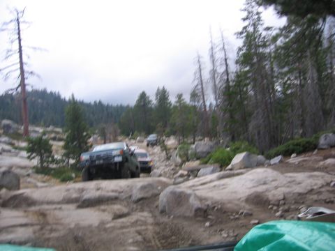 1989 Ford Bronco - Multiple 4x4 vehicles are seen traversing a difficult, rocky trail in a wooded area.