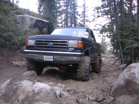 1989 Ford Bronco - A lifted black Ford Bronco navigates a challenging off-road trail with large rocks and trees.
