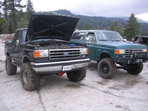 1989 Ford Bronco - Two Ford Broncos are parked on a rocky surface with trees and mountains in the background. The black Bronco has its hood open.
