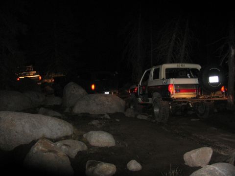 1989 Ford Bronco - Several off-road vehicles navigate a rocky path in a wooded area after dark.
