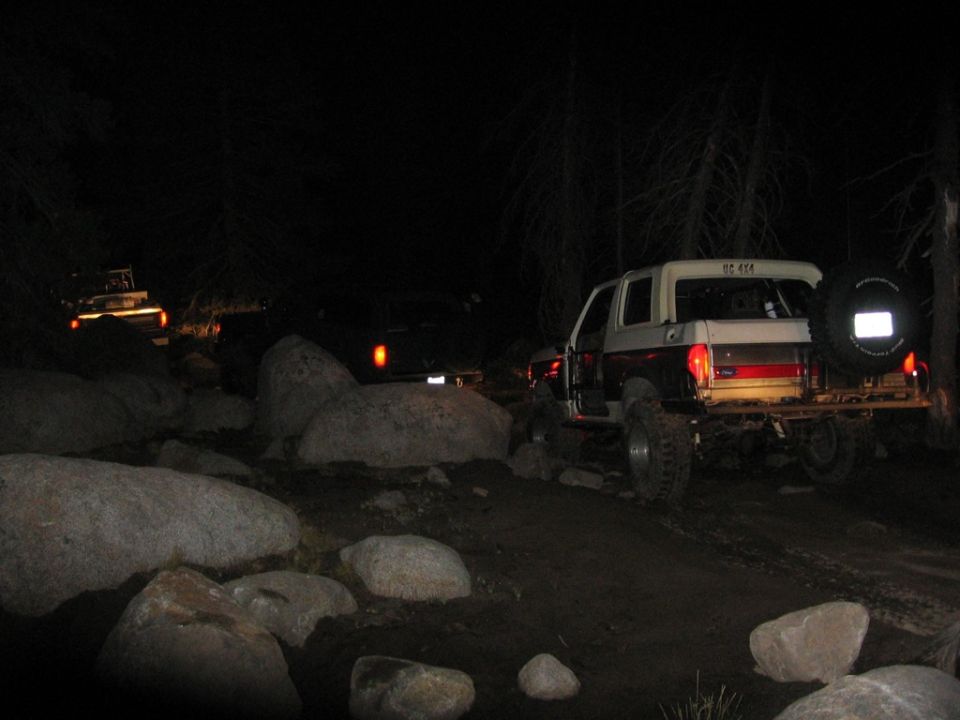 Two off-road vehicles driving on a rocky trail at night. The lead vehicle is a 1989 Ford Bronco