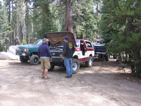 1989 Ford Bronco - Two men stand by a red and white 1989 Ford Bronco with its hood open, while a green Ford F-Series truck is parked nearby in a wooded area.