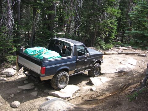 1989 Ford Bronco - A Ford Bronco navigates a challenging, rocky off-road trail surrounded by trees.