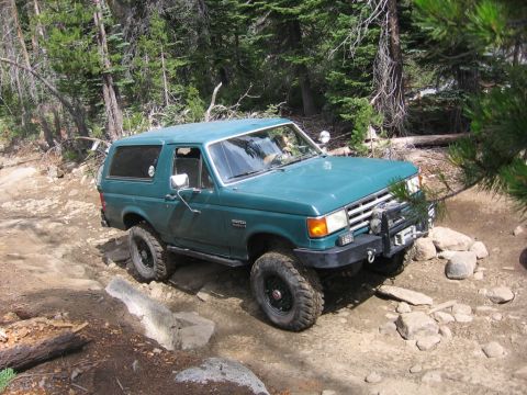 1989 Ford Bronco - Green 1989 Ford Bronco tackling a challenging rocky trail in a wooded area.